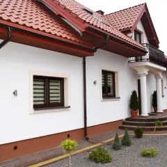 Biały dom z brązowym dachem i rynnami. Widać schody, okna i ozdobne krzewy. Cokół domu jest brązowy.