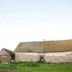 Stary budynek gospodarczy z częścią mieszkalną, zniszczony tynk, zardzewiały dach, zarośnięty teren.