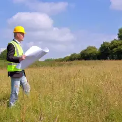 Inżynier w żółtym kasku i kamizelce odblaskowej analizuje plany działki, zastanawiając się, jak odlesić działkę i rozpocząć budowę.