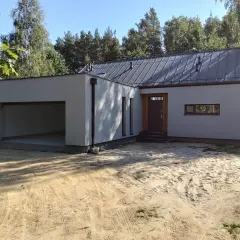 Nowoczesny garaż z płaskim dachem, otwarty wjazd, drzwi i wąskie okno. Otoczenie piaszczyste, drzewa w tle.