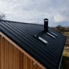 Czarna blacha na dach dwuspadowy, z widocznymi oknami dachowymi i kominem. Elewacja z drewna.