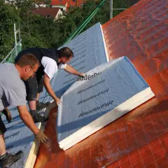 Dwóch budowlańców układa płyty izolacyjne na dachu. To kluczowy etap ocieplenia dachu od wewnątrz, zapewniający komfort termiczny.