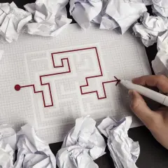 Ręka rysuje drogę przez labirynt na kratkowanym papierze, otoczona zgniecionymi kartkami. To symbol przeniesienia warunków zabudowy, szukania rozwiązania.