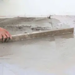 Ręka wyrównuje mokry beton poziomnicą. Powierzchnia jest w trakcie prac, a w tle widać elementy konstrukcyjne.