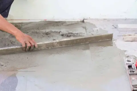 Ręka wyrównuje mokry beton poziomnicą. Powierzchnia jest w trakcie prac, a w tle widać elementy konstrukcyjne.