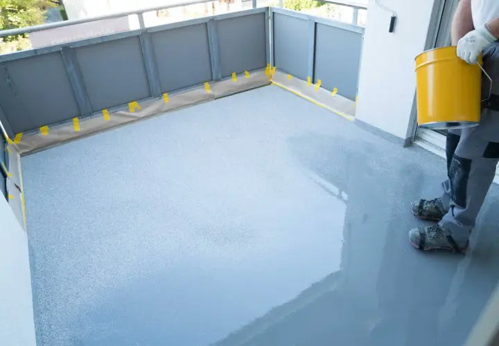 Pracownik wykonuje hydroizolację balkonu, nakładając szarą masę na zabezpieczoną taśmą posadzkę.