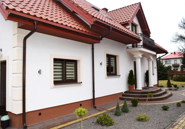 Biały dom z brązowym dachem i rynnami. Widać schody, okna i ozdobne krzewy. Cokół domu jest brązowy.