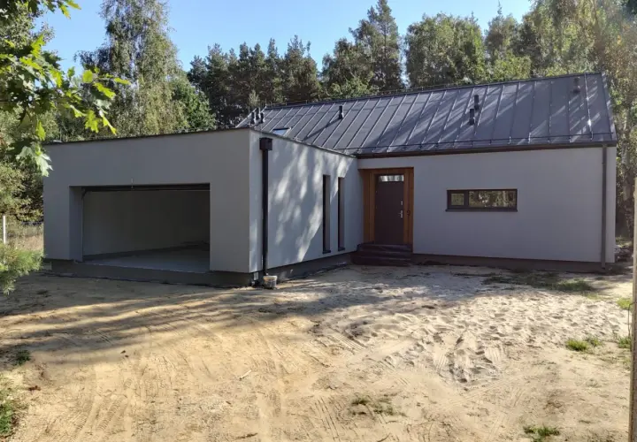 Nowoczesny garaż z płaskim dachem, otwarty wjazd, drzwi i wąskie okno. Otoczenie piaszczyste, drzewa w tle.