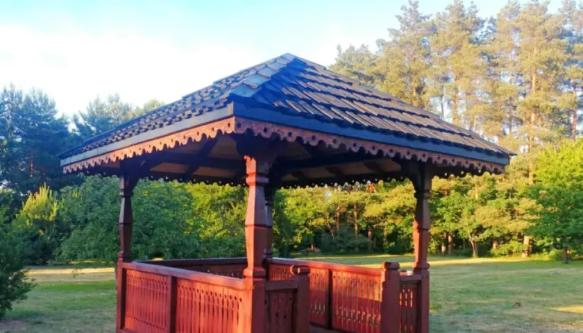 Elegancka altana z bali projekt, z rzeźbionymi elementami i drewnianym dachem, otoczona zielenią.