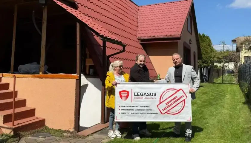 Trzy osoby trzymają baner "Budynek zalegalizowany". Pomagają w procesie jak zrobić odbiór domu po 20 latach.