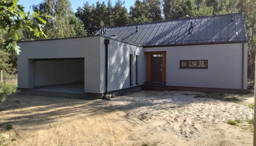 Nowoczesny garaż z płaskim dachem, otwarty wjazd, drzwi i wąskie okno. Otoczenie piaszczyste, drzewa w tle.