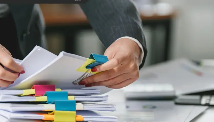 Ręce sortują dokumenty, wśród nich pozwolenie na budowę. Kolorowe spinacze porządkują stosy papierów.