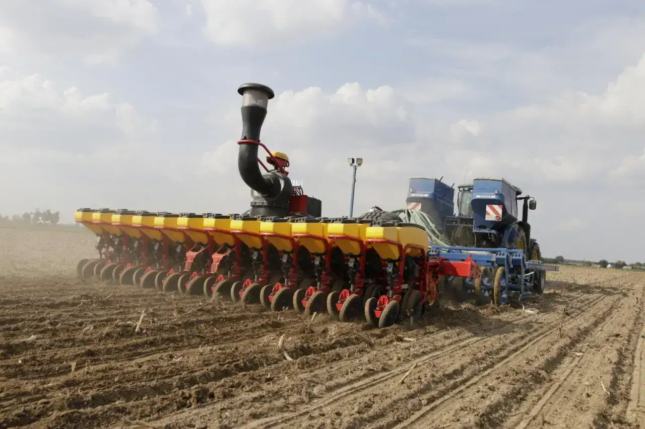 Siewnik punktowy z żółtymi zbiornikami wysiewa nasiona na polu, za nim widać traktor.