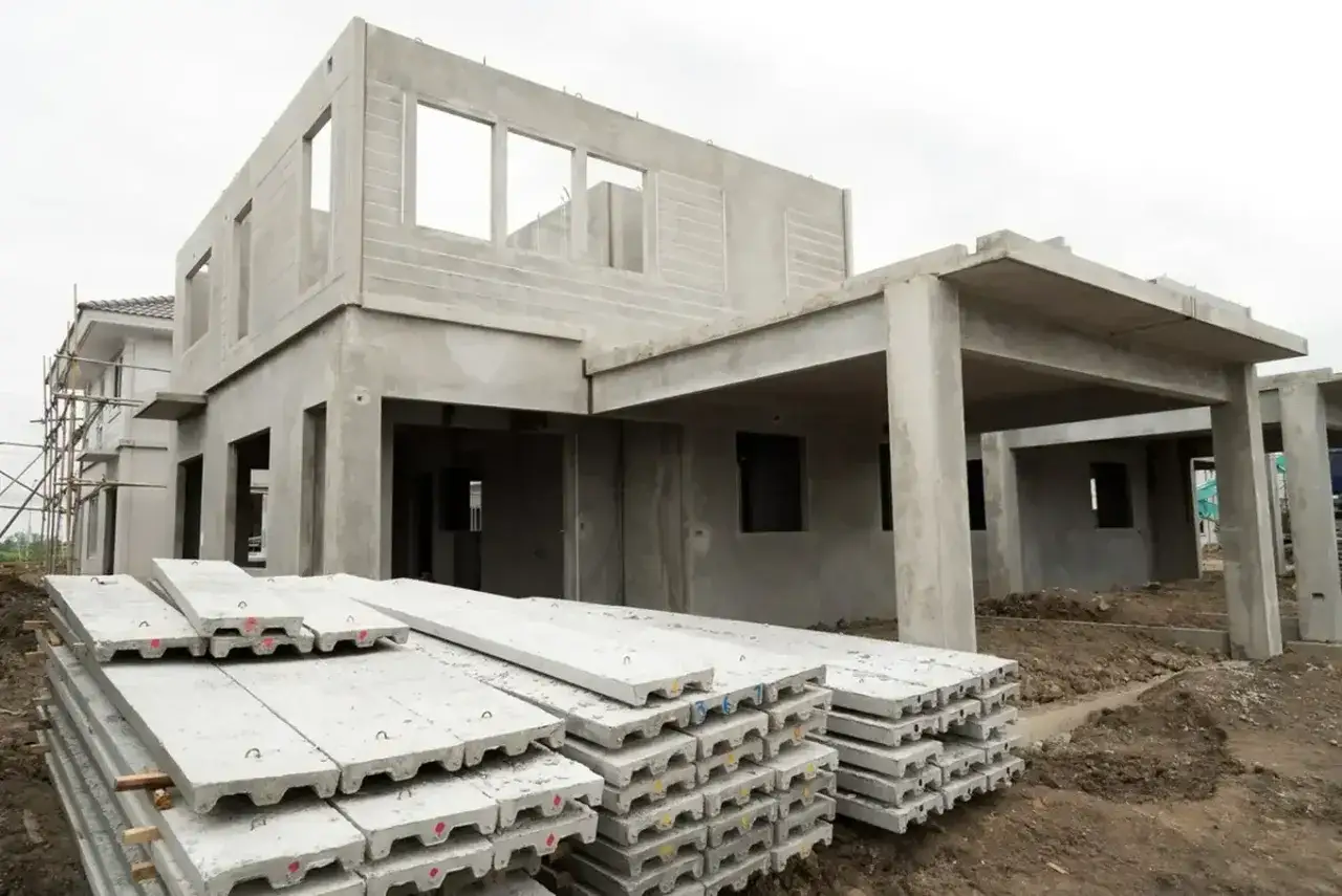 Budowa domu z płyt betonowych w toku. Na pierwszym planie stosy płyt, w tle surowa konstrukcja budynku z otworami okiennymi i drzwiami.