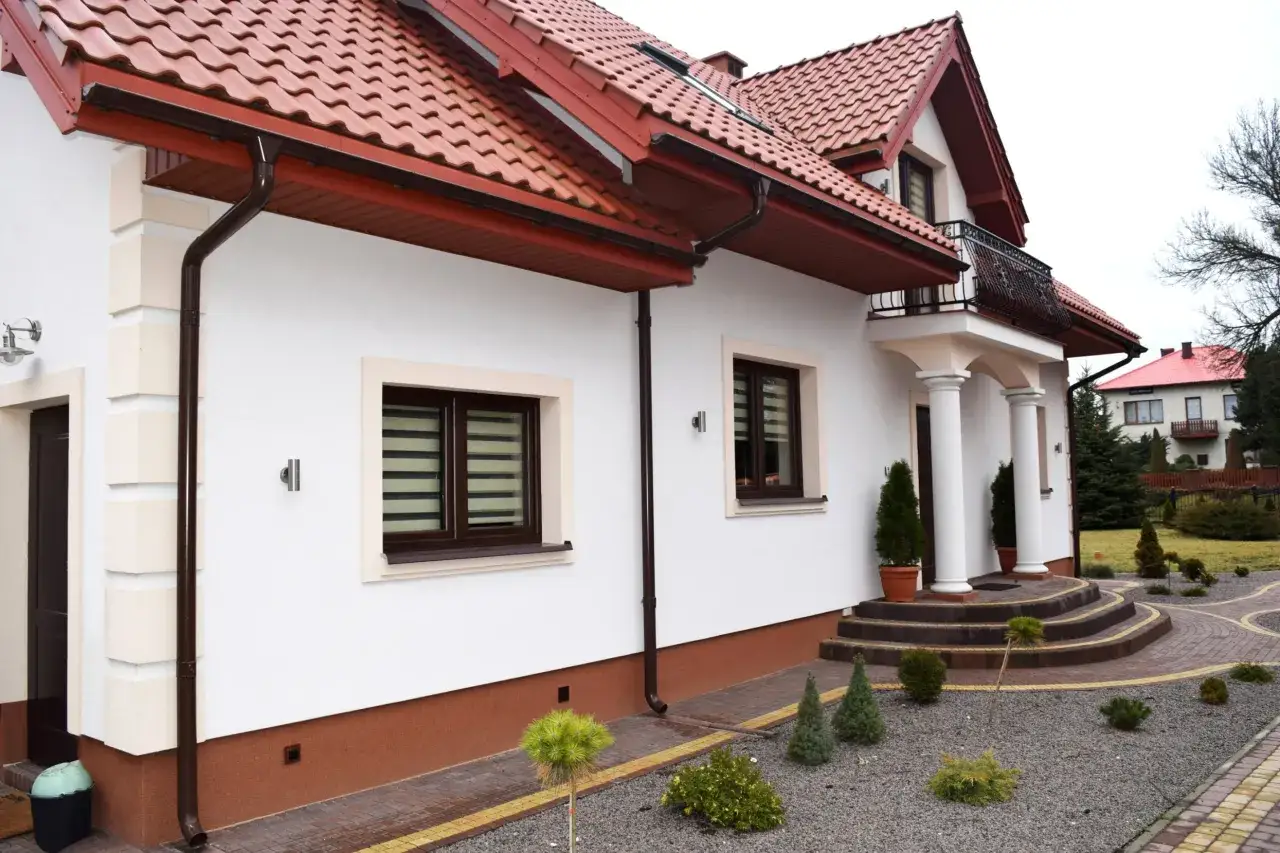 Biały dom z brązowym dachem i rynnami. Widać cokół, schody i wejście.
