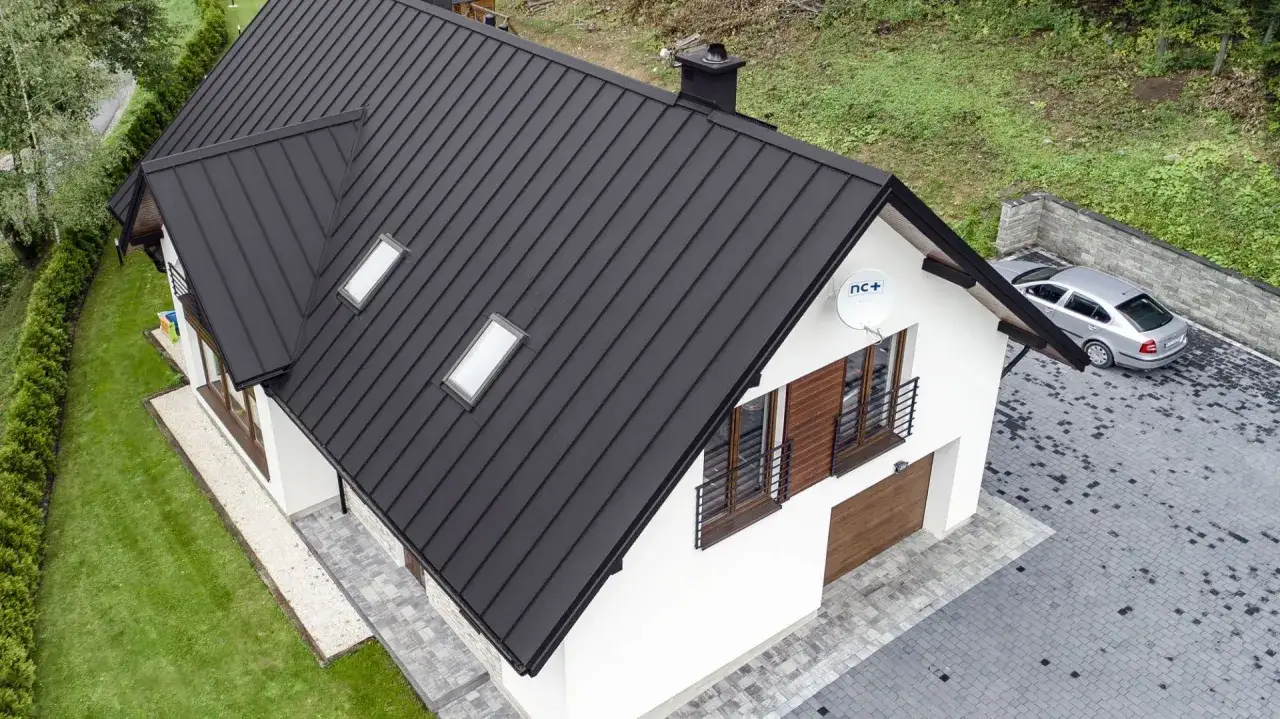Nowoczesny dom z czarną blachą na rabek na dachu. Obok podjazd z kostki brukowej i srebrny samochód.